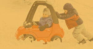One young student pushes another young student in a toy car through the snow.