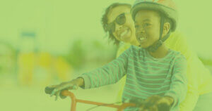Young student smiles as his mother pushes him on his bike with his helmet.